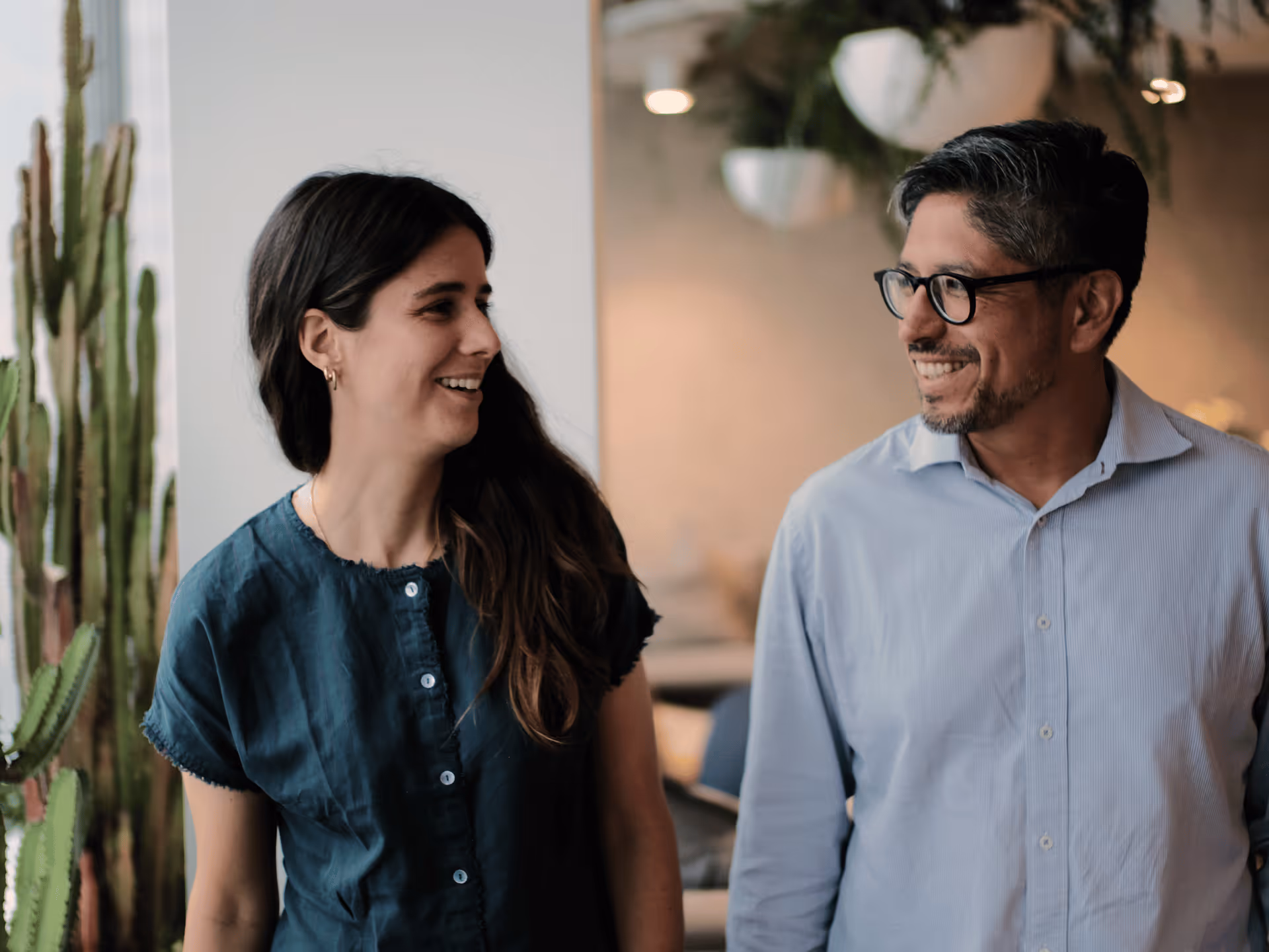 Smiling man and woman walking indoors near a window with tall green plants.