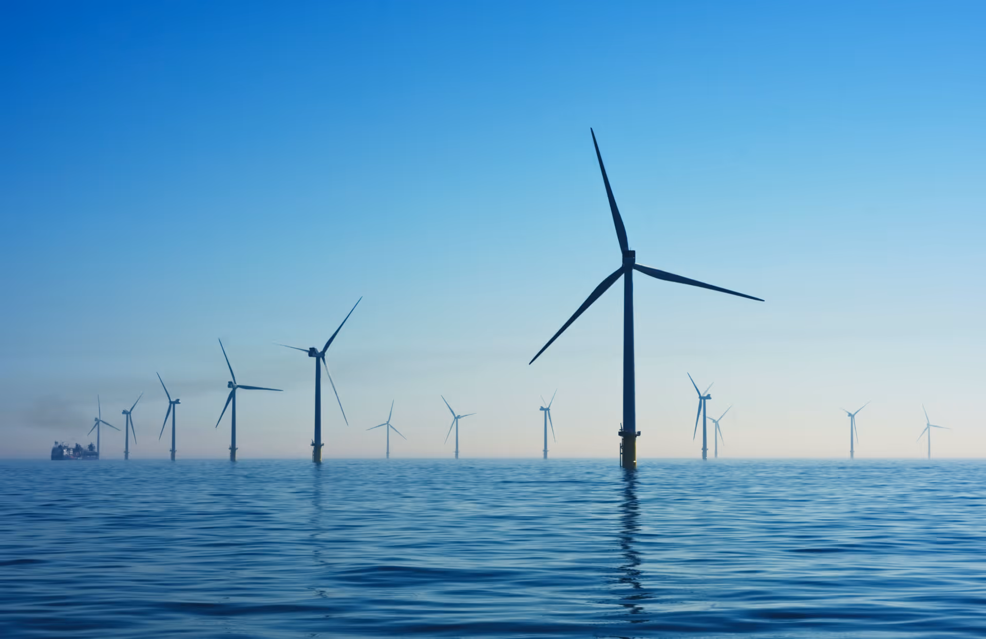 Offshore wind turbines standing in the sea with a ship in the background under a clear blue sky.