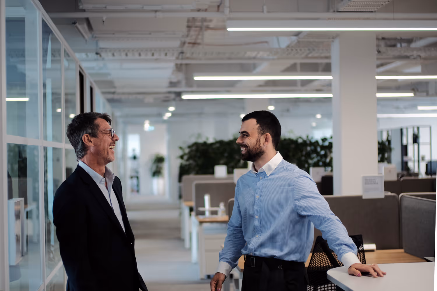 Two men smiling and talking in a modern office with desks and glass partitions.