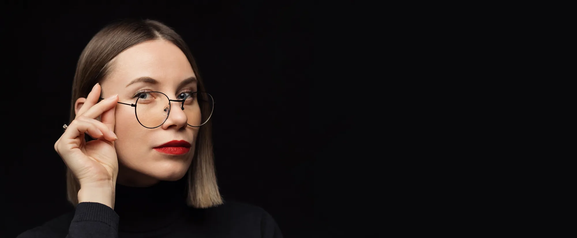Woman with straight blond hair, red lipstick, and round glasses on a black background.