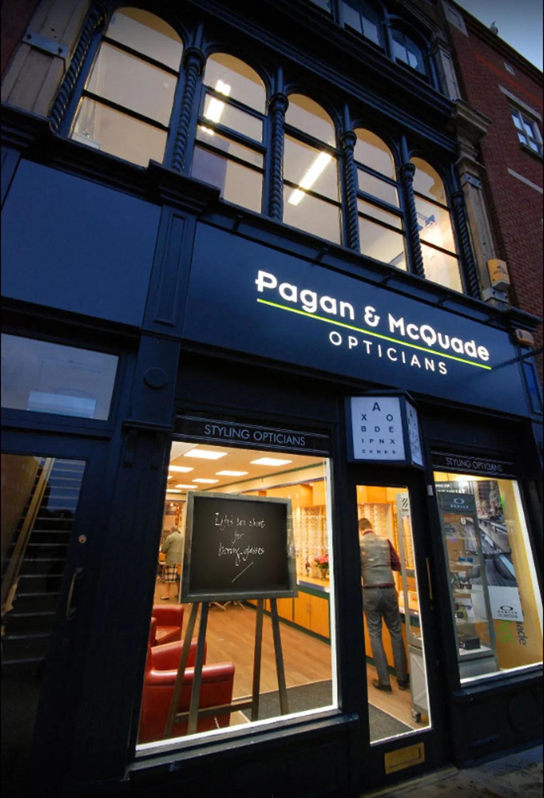 Pagan & McQuade Opticians storefront with blackboard sign and person inside at dusk.