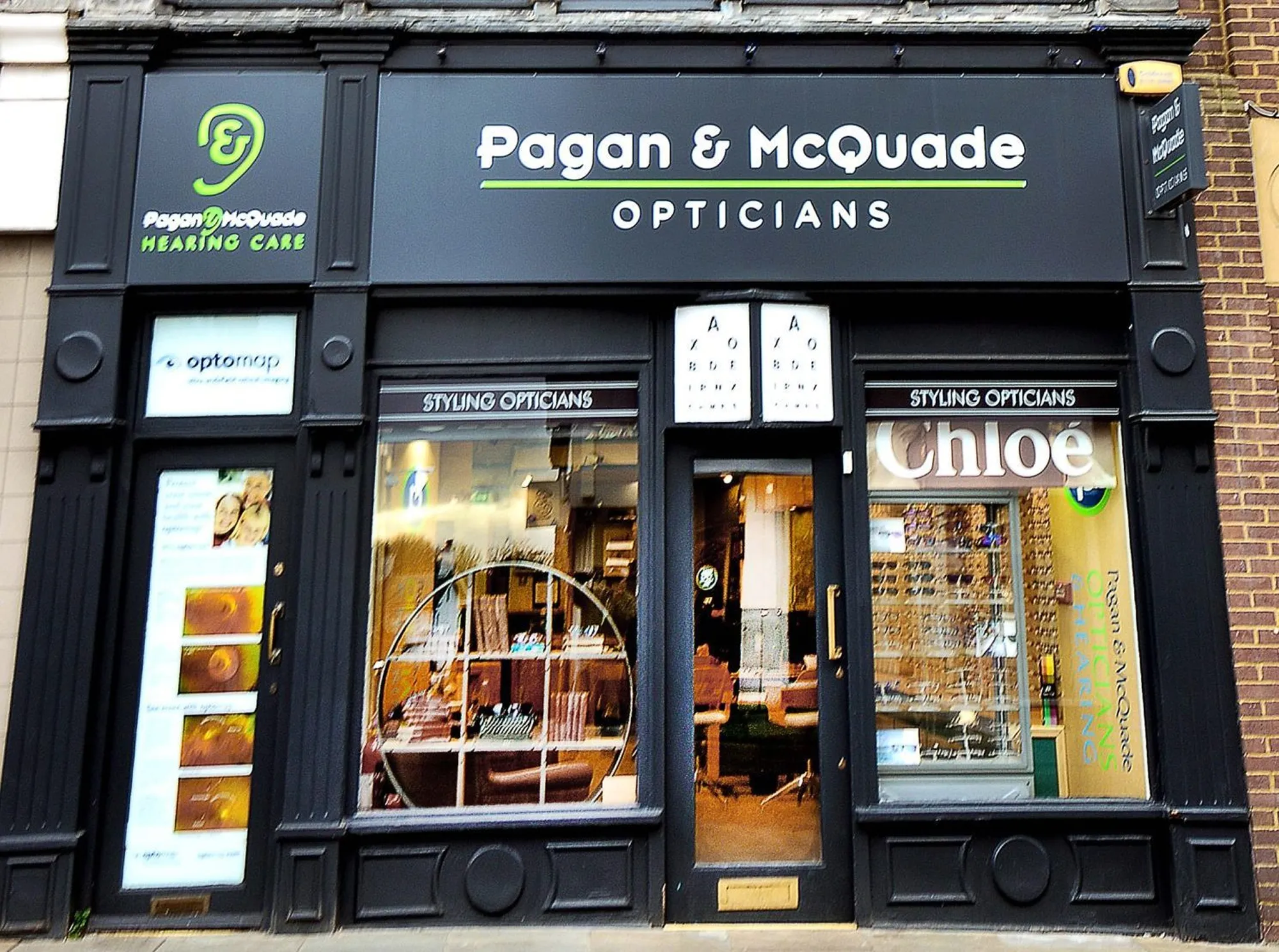 Storefront of Pagan & McQuade Opticians and Hearing Care with eye chart and glasses displays.
