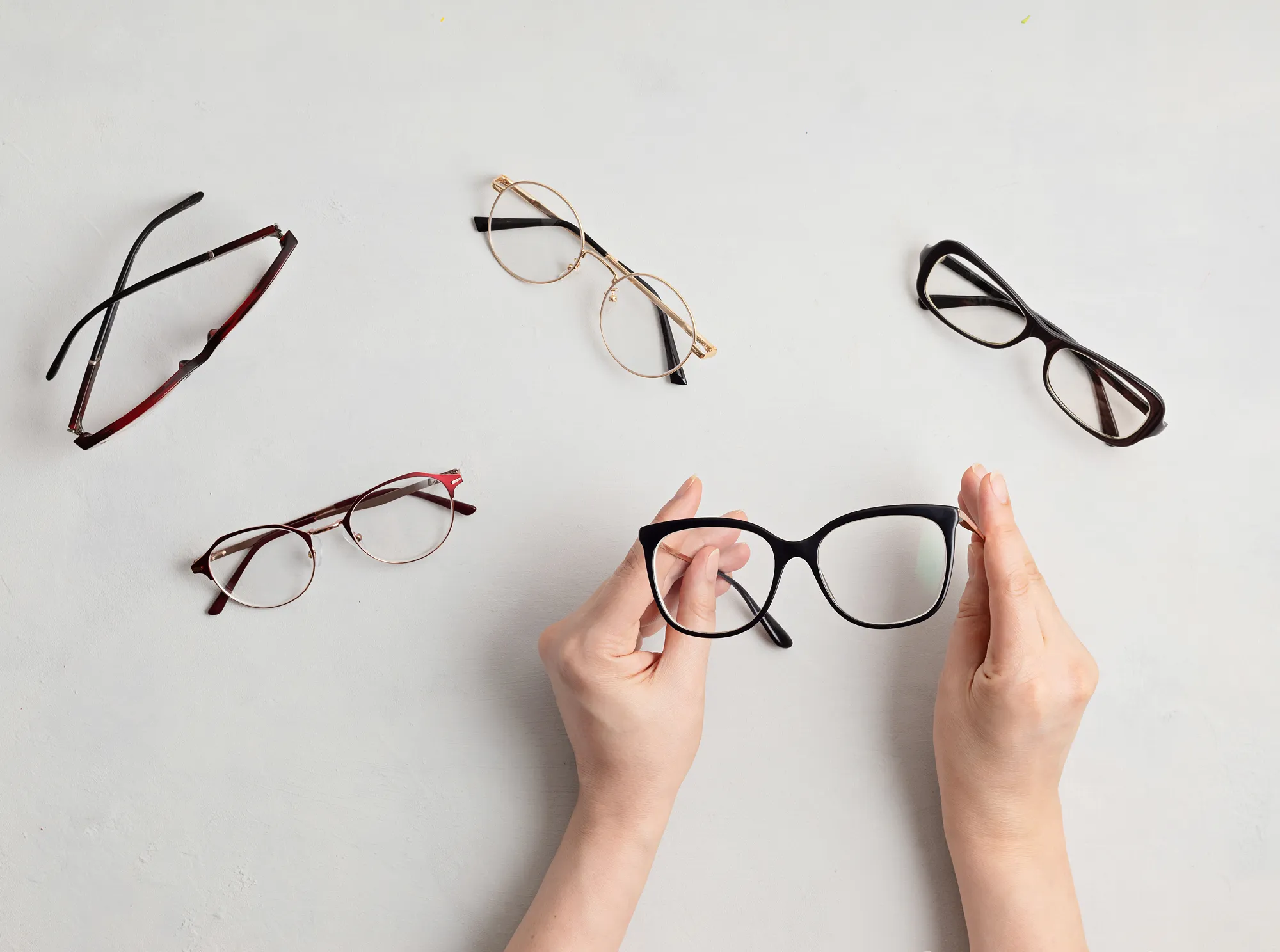 Hands holding a pair of black eyeglasses with four other pairs on a white surface.