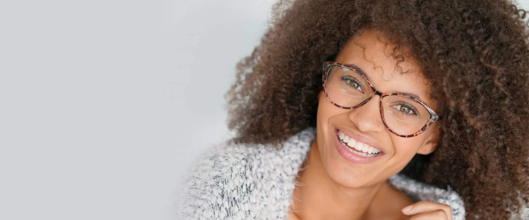 Smiling woman with curly hair wearing tortoiseshell glasses and a gray sweater against gray background.