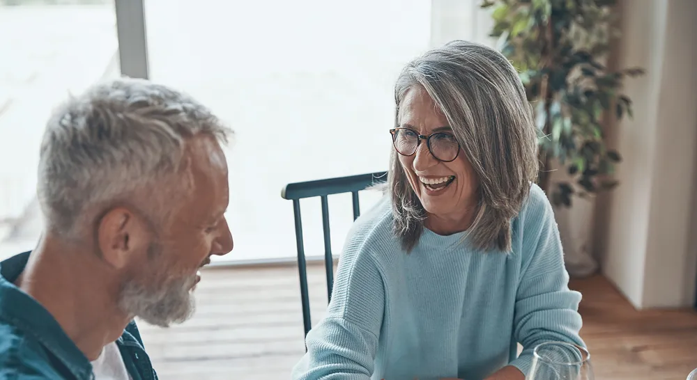 Older woman with glasses laughing and talking with an older man in a bright room.