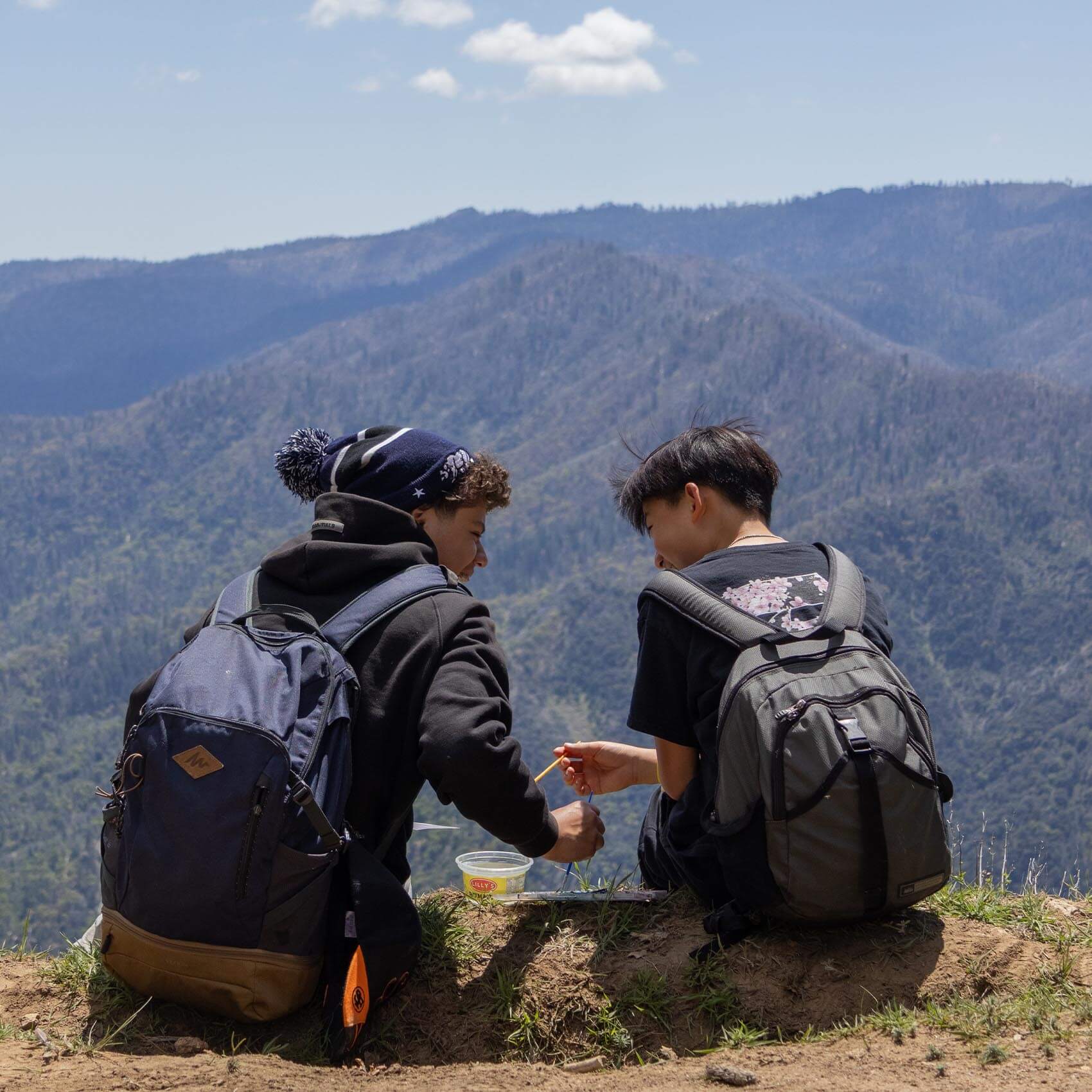 Two young hikers with large backpacks sit on a dirt ridge overlooking a vast, hazy mountain range under a clear blue sky. They appear to be sharing a snack or looking at something small together.