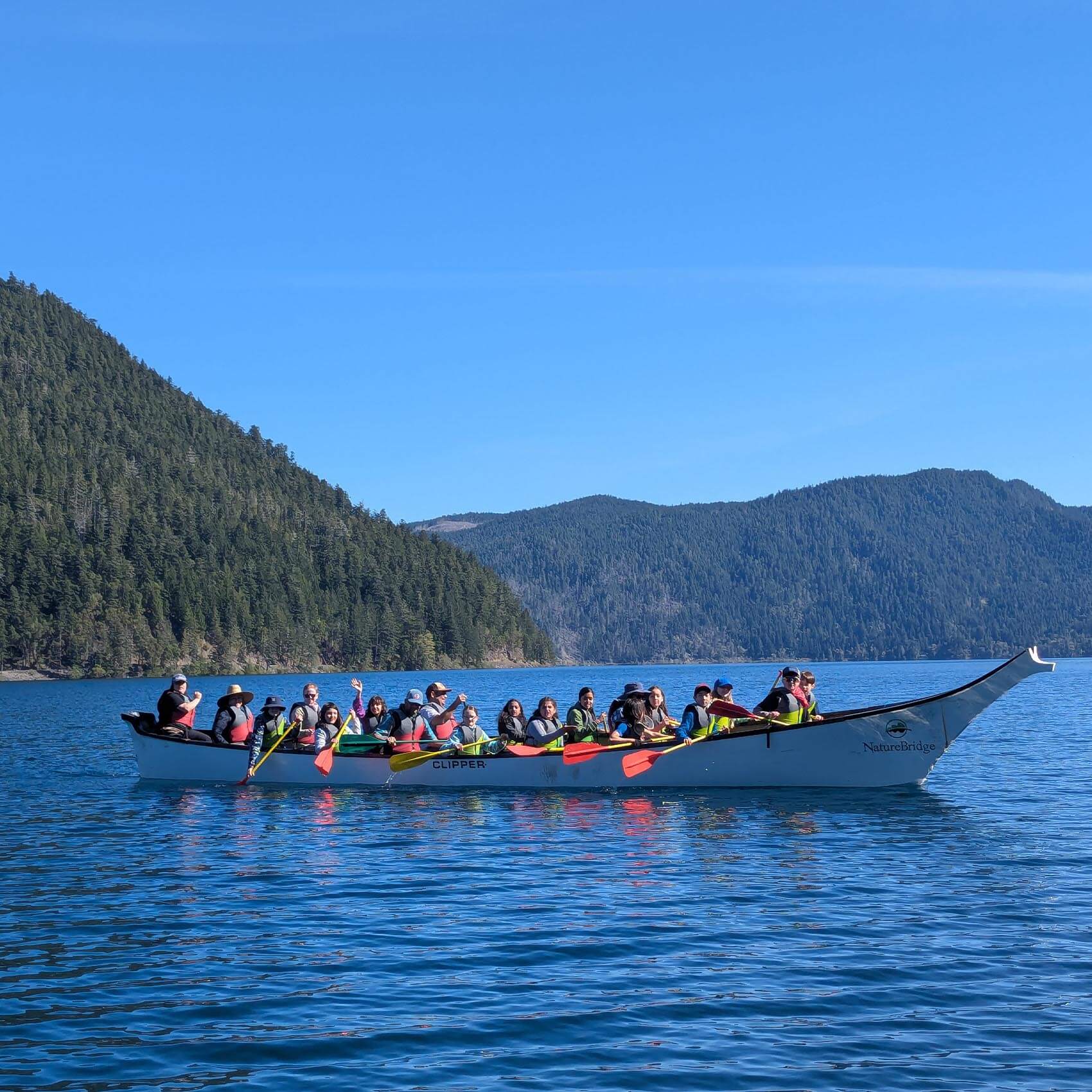 A long white "NatureBridge" canoe filled with about twenty people wearing life jackets, paddling across a deep blue lake. Large, tree-covered hills rise steeply from the water's edge in the background.