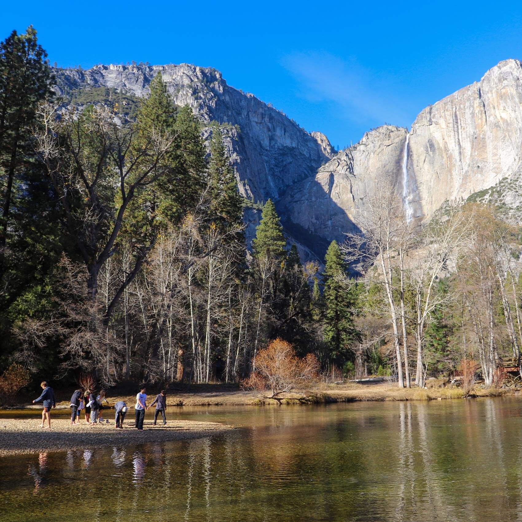 A wide landscape of Yosemite Valley featuring a high waterfall cascading down a sheer granite cliff. In the foreground, a small group of people stands on a rocky riverbank next to a calm reflection of the mountains in the water.