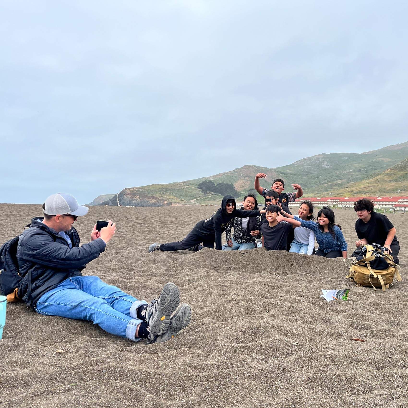 A man sits on a sandy beach using his smartphone to take a photo of a group of kids who are buried up to their waists in the sand, laughing and making poses.