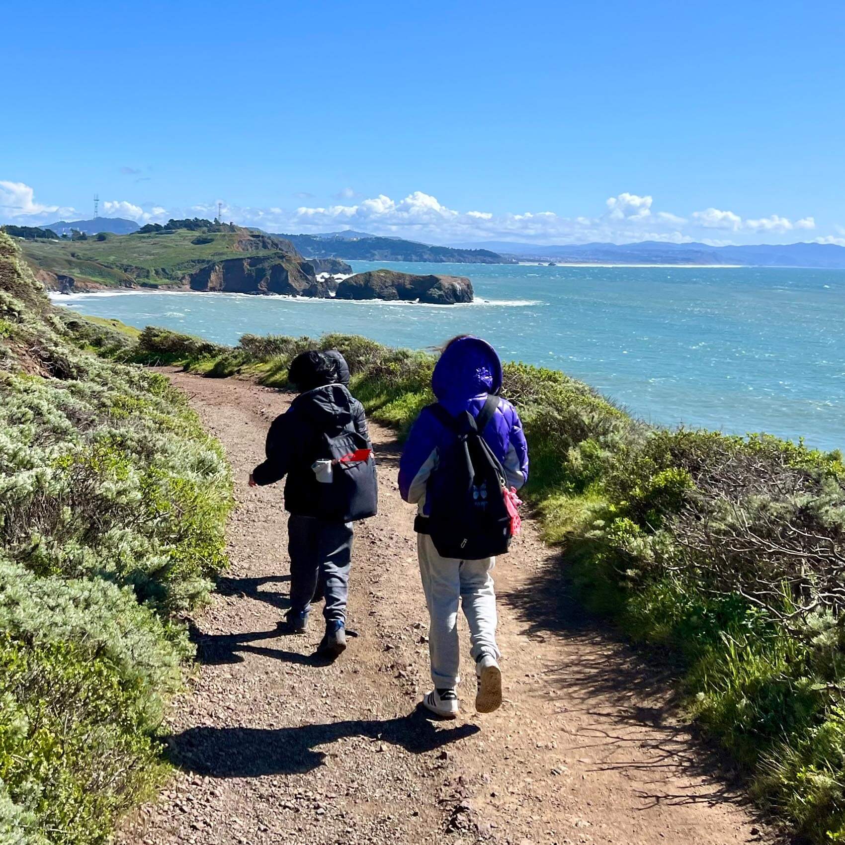 Two backpackers walk away from the camera along a dirt trail on a grassy coastal bluff. The bright blue ocean and a rugged coastline with rocky outcrops are visible to the right.
