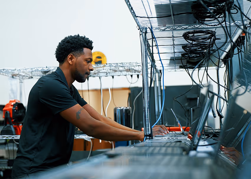 Man with black hair and a black shirt working on a laptop in a tech or server room with multiple laptops and cables.