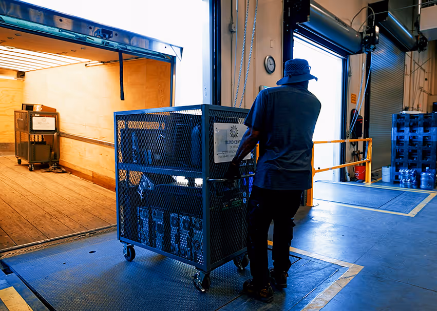 Worker wearing a hat pushing a large metal cart loaded with boxes into an open delivery truck inside a warehouse.