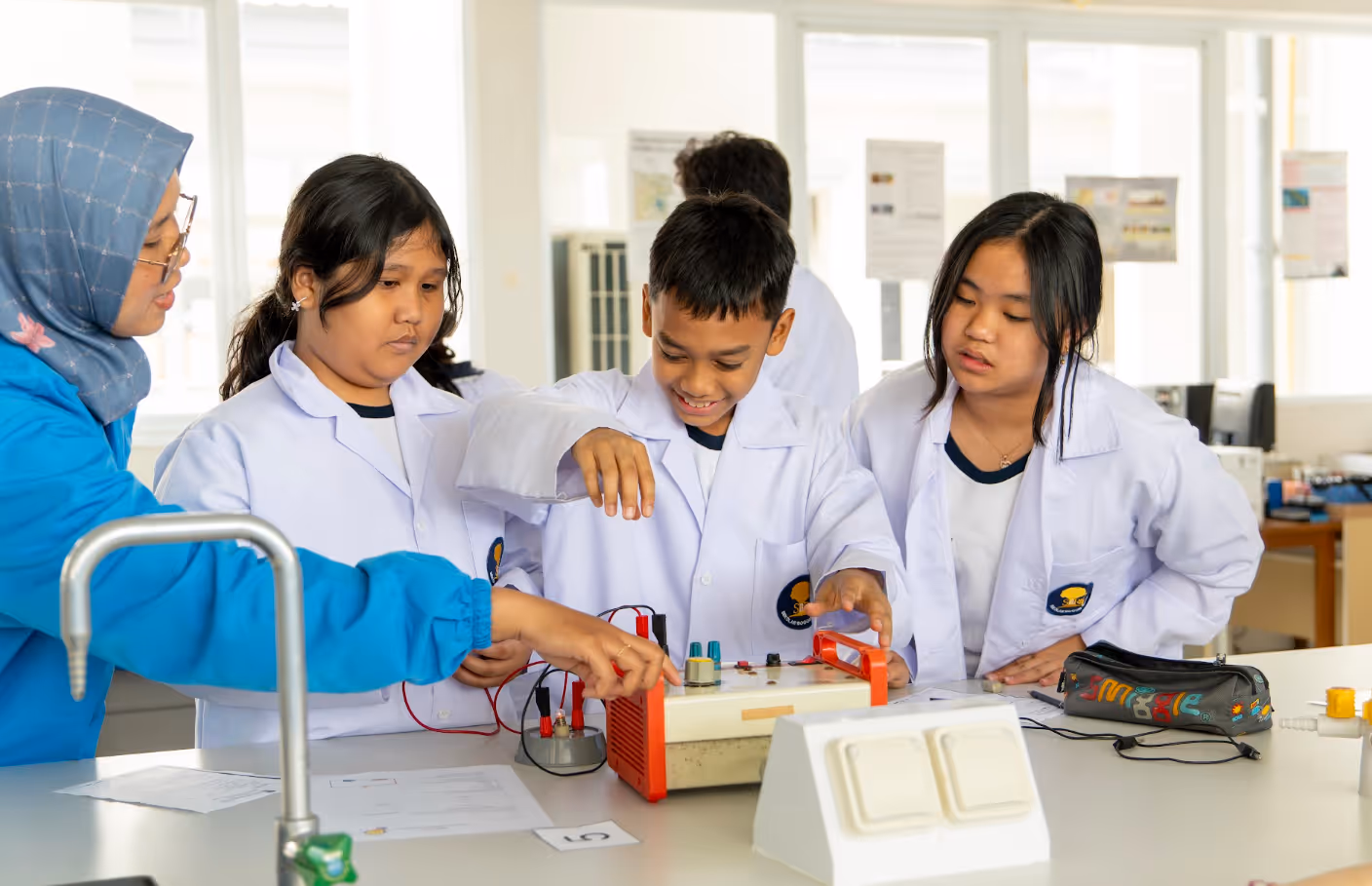 Teacher guiding three Sekolah Bogor Raya students in white lab coats using electronic lab equipment in a bright science classroom.
