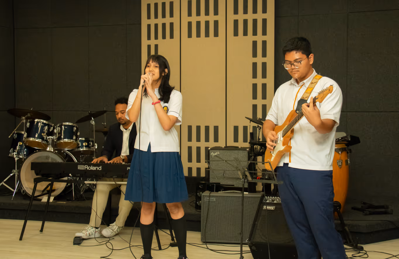 Students of Sekolah Bogor Raya singing with microphone, playing electric guitar, and teacher playing keyboard on stage.