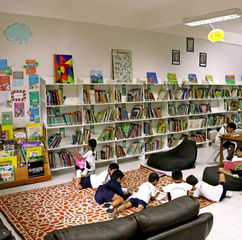 Sekolah Bogor Raya's Primary School Library. Children in school uniforms reading and writing on a carpet and bean bags in a library with bookshelves.