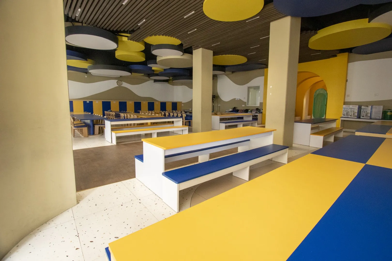 Sekolah Bogor Raya's Primary School Cafeteria. Indoor seating area with yellow and blue benches, tables, and circular ceiling panels in a modern space.