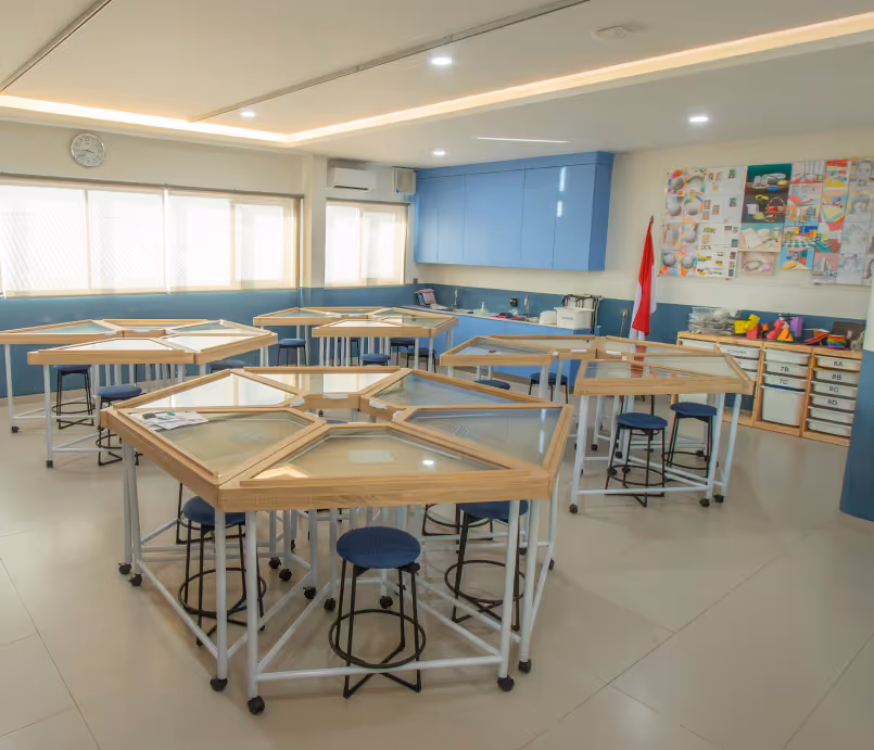 Sekolah Bogor Raya's Secondary School Art Room. Bright classroom with hexagonal tables, blue stools, storage shelves, and a wall clock.