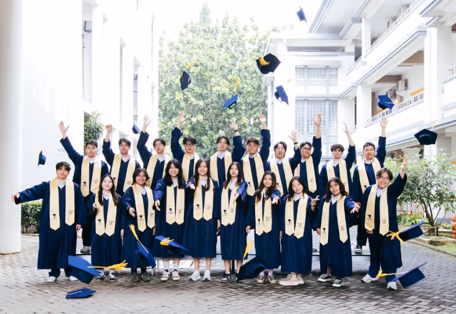 Sekolah Bogor Raya students in blue gowns and gold stoles throwing caps during graduation outside school building. Graduating class of IB Diploma programme.