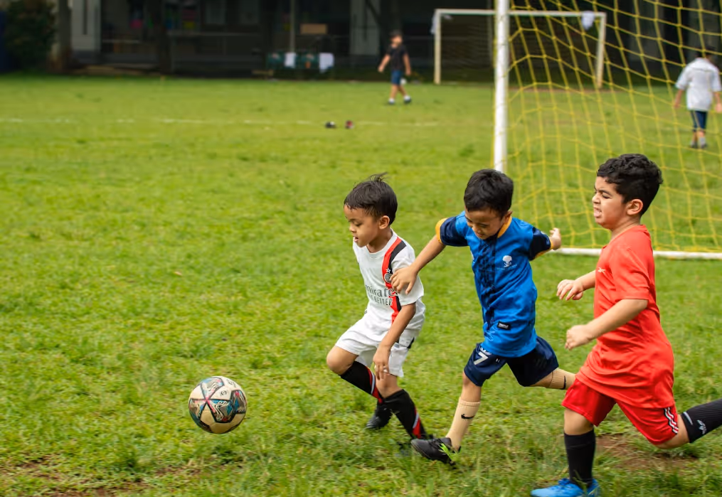 Soccer Field of Sekolah Bogor Raya. Three young boys in sportswear chasing a soccer ball on a grassy field near a goalpost.