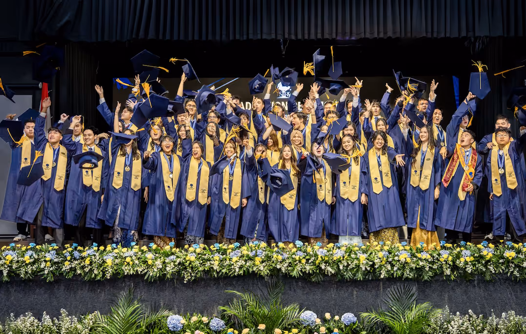Group of Sekolah Bogor Raya graduates in blue gowns and gold stoles throwing caps on stage with flowers below.