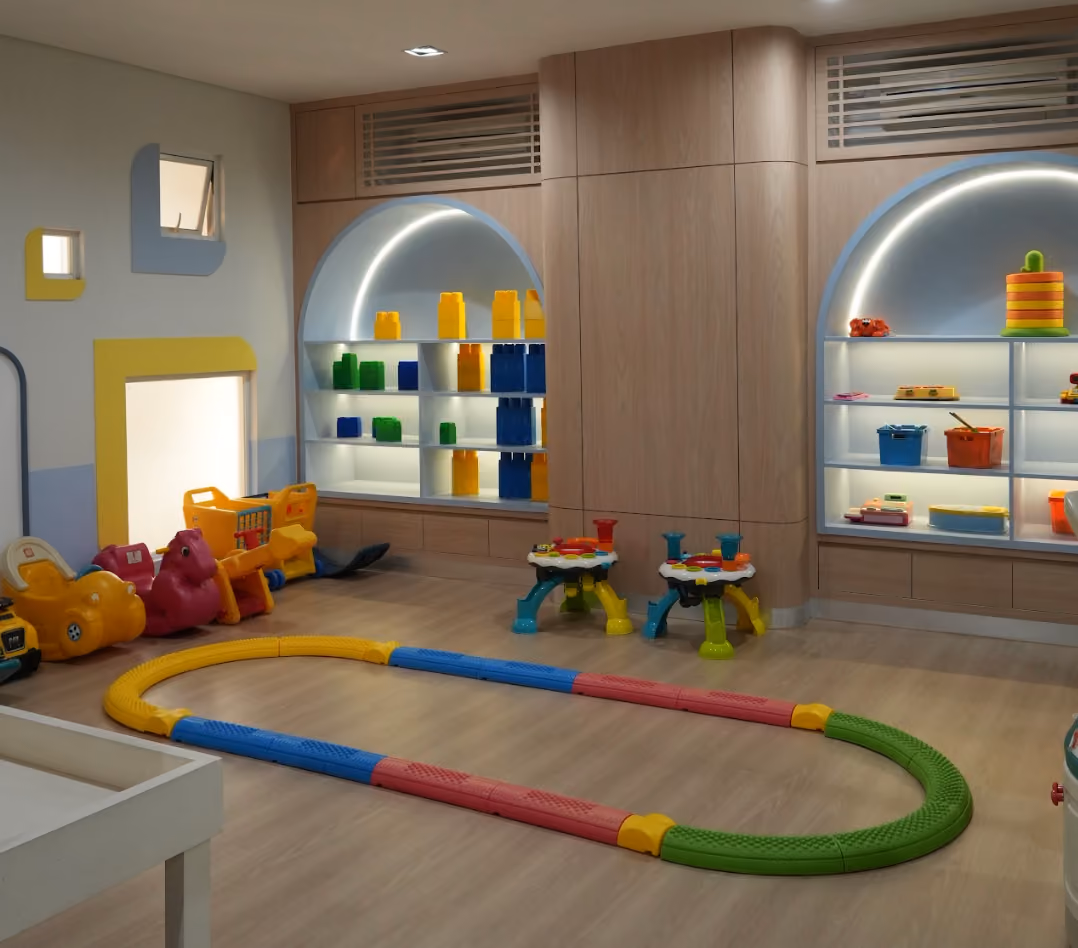 Indoor playgroup and kindergarten sensory room with wooden flooring and modern shelves displaying colorful large building blocks and toys, including yellow, green, and blue pieces. The room has two small activity tables with multiple colors and a low track made of interlocking pieces in yellow, blue, red, and green on the floor. On the left, various ride-on toys in orange, red, and yellow are lined up near a wall with small windows framed in blue and yellow. The walls and lighting create a cozy and organized play space.