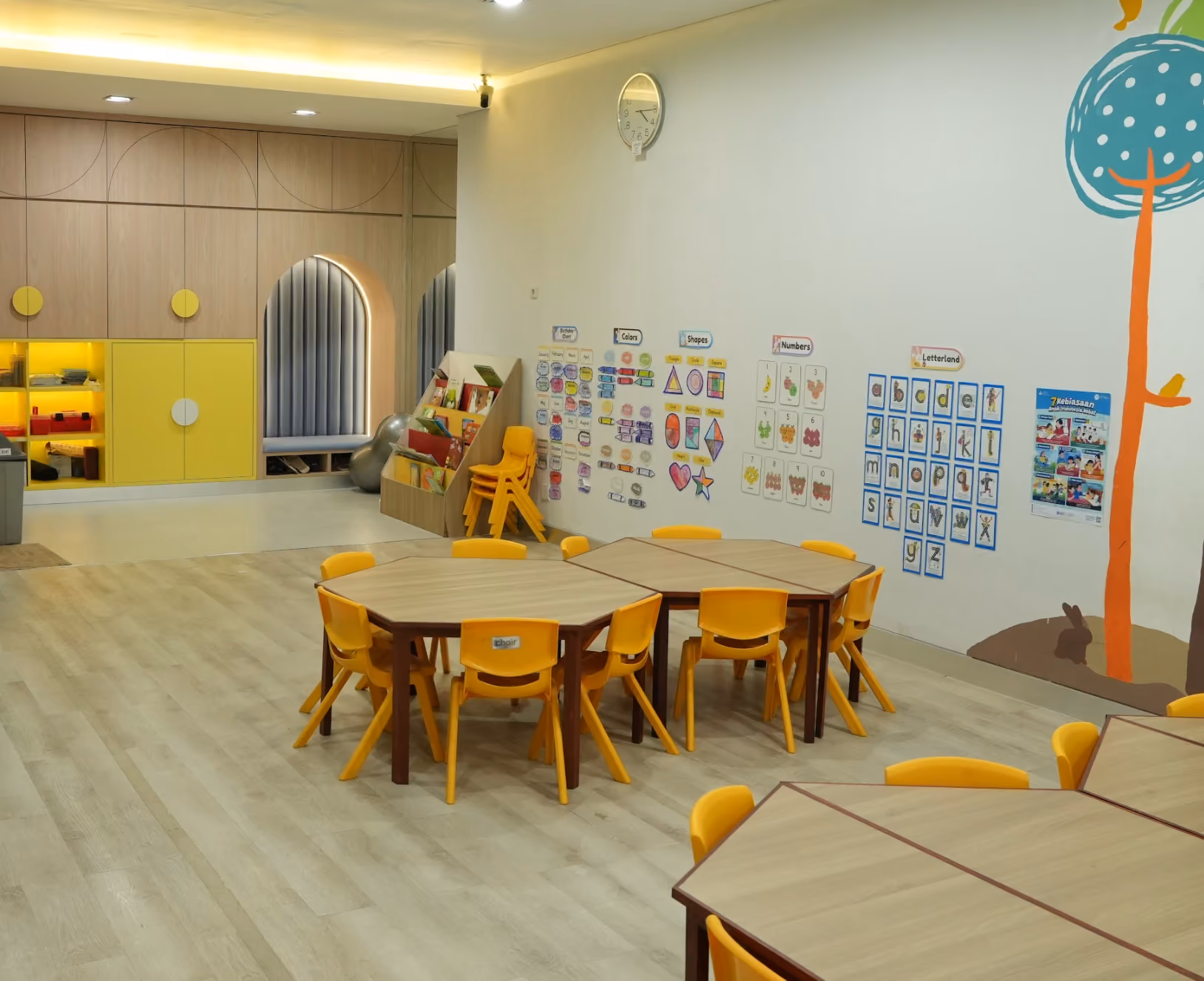 A brightly lit playgroup and kindergarten classroom with yellow kids' chairs around hexagonal wooden tables, educational posters on the wall showing colors, shapes, numbers, and letters, and a colorful tree mural with a rabbit at its base. There is a book rack and additional stacked chairs near wooden storage cabinets with yellow doors, and a clock on the wall shows the time as 9:10.