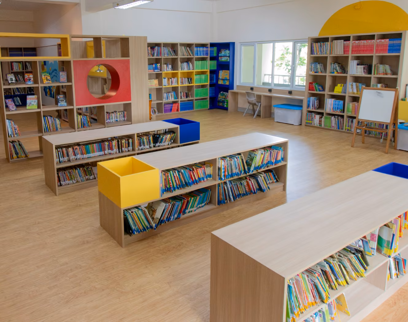Primary School library with wooden bookshelves filled with colorful books, some shelves featuring yellow and blue cubbies, a window with chairs and desks below, and a whiteboard on an easel.