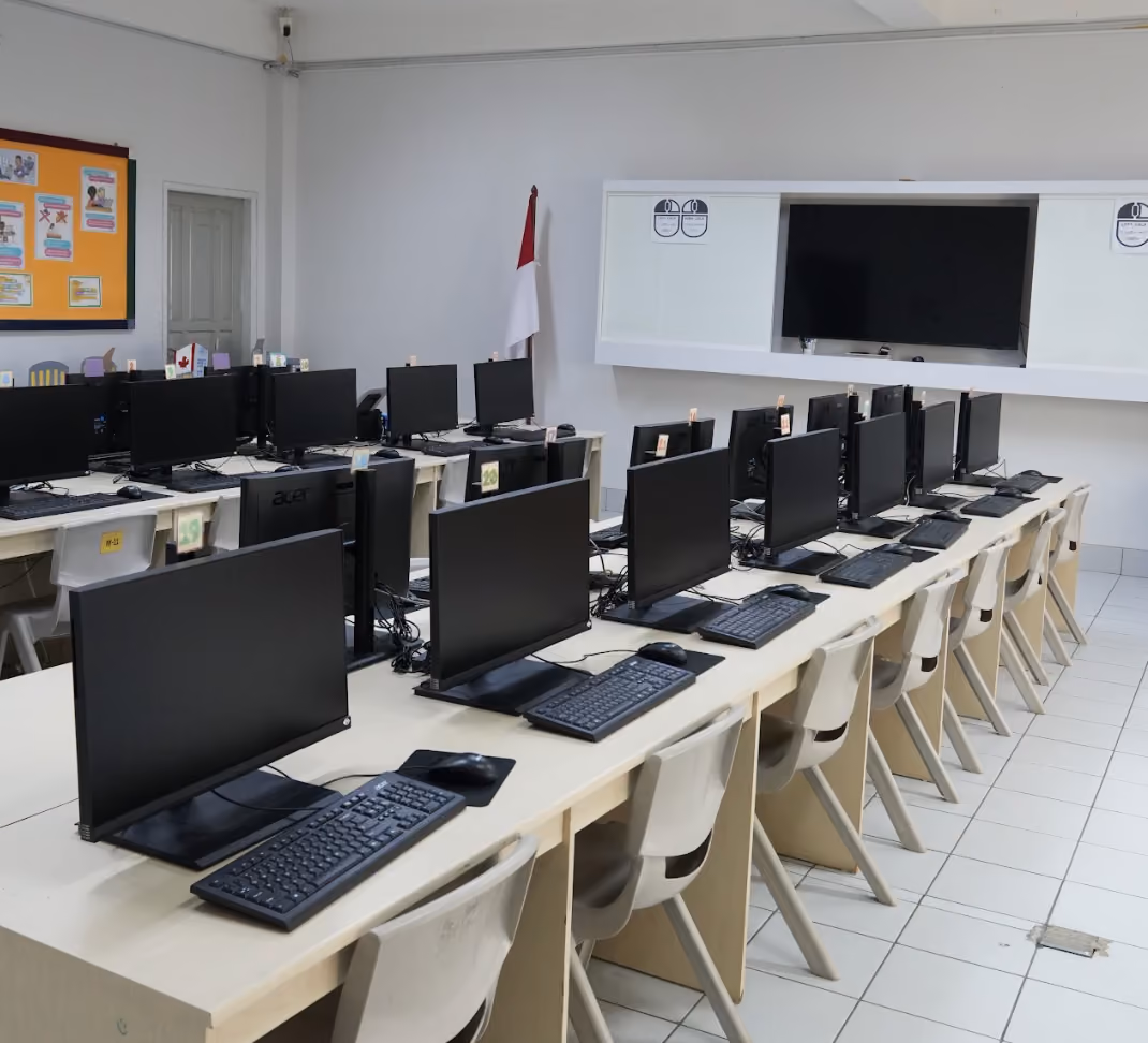 Primary school computer lab with multiple rows of desks, each equipped with a black monitor, keyboard, and mouse. Beige chairs are neatly arranged at each station. A large screen is mounted on the white wall at the front of the room, and an Indonesian flag stands in the corner. A bulletin board with colorful notices is visible on the left wall.