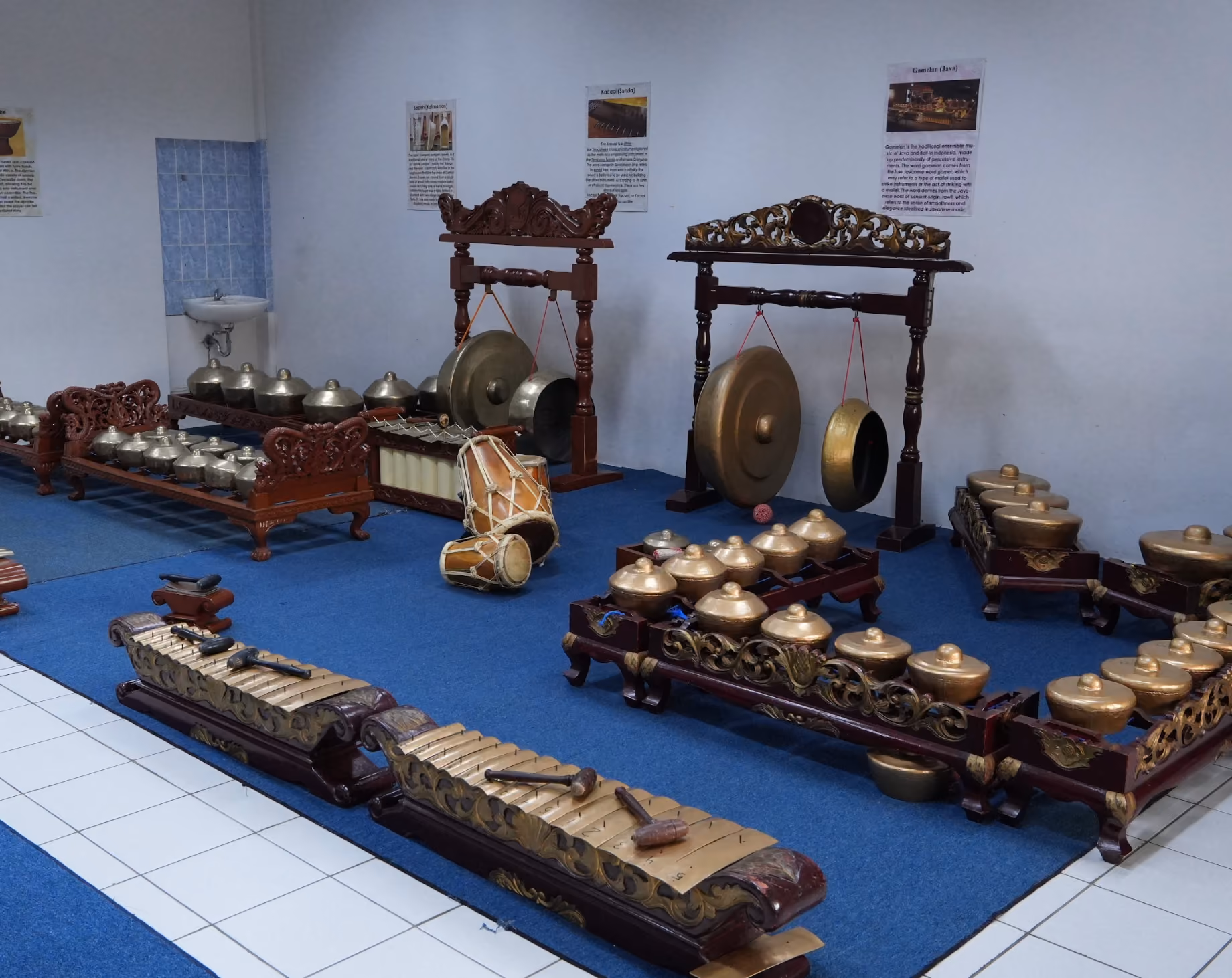Primary School's Traditional Music Room displaying a traditional Indonesian gamelan ensemble with various bronze gongs, metallophones, drums, and wooden frames on a blue carpet against a white wall with informational posters.
