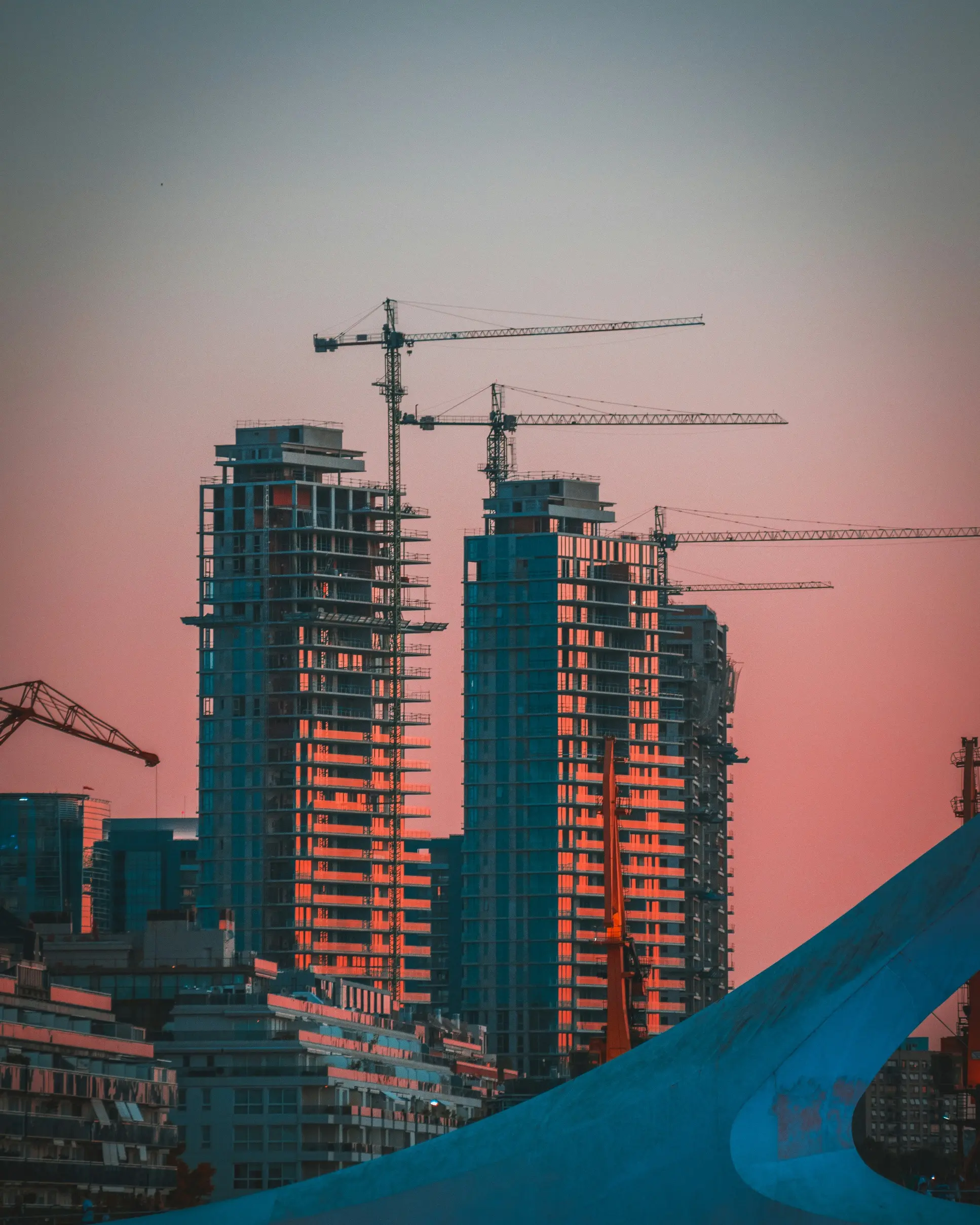 Two high-rise buildings under construction with cranes against a pink and blue sky at sunset.