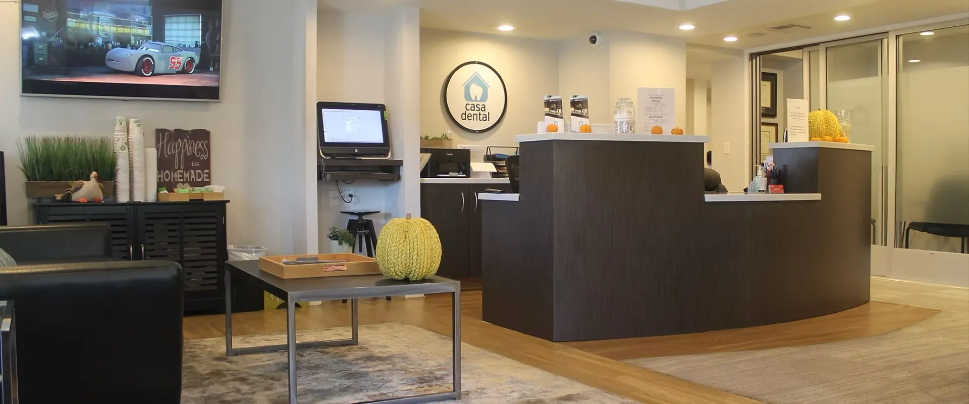 Modern dental office waiting room with black chairs, a coffee station, TV showing animation, and a reception desk with Casa Dental logo.
