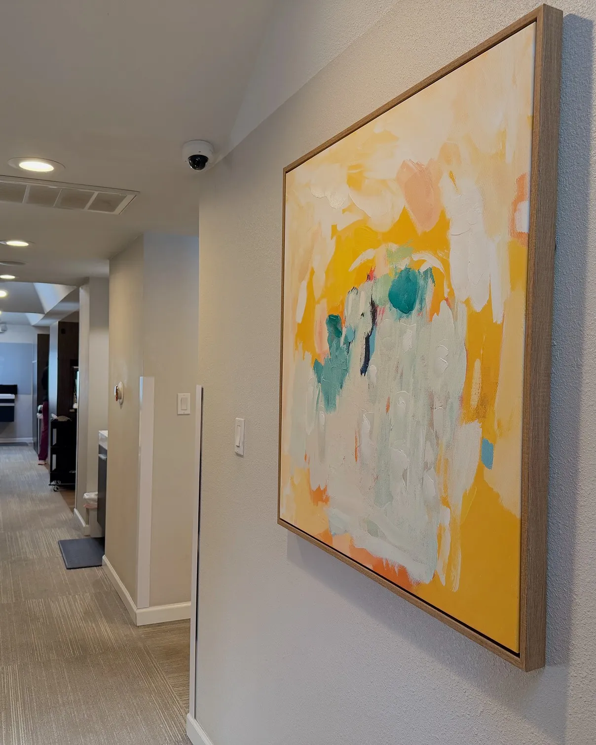 Abstract painting with yellow, white, and teal brushstrokes hanging on a beige hallway wall with carpeted floor and ceiling lights.