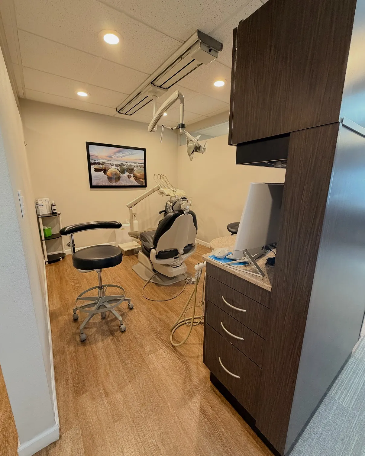 Dental office room with a dental chair, dentist stool, overhead dental light, wooden flooring, and a framed picture of rocks on a beach wall.