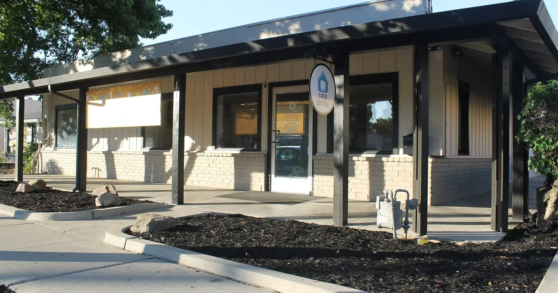 Exterior view of Casa Dental clinic with beige brick walls, a white door, and a hanging round sign displaying the clinic's blue and white logo.