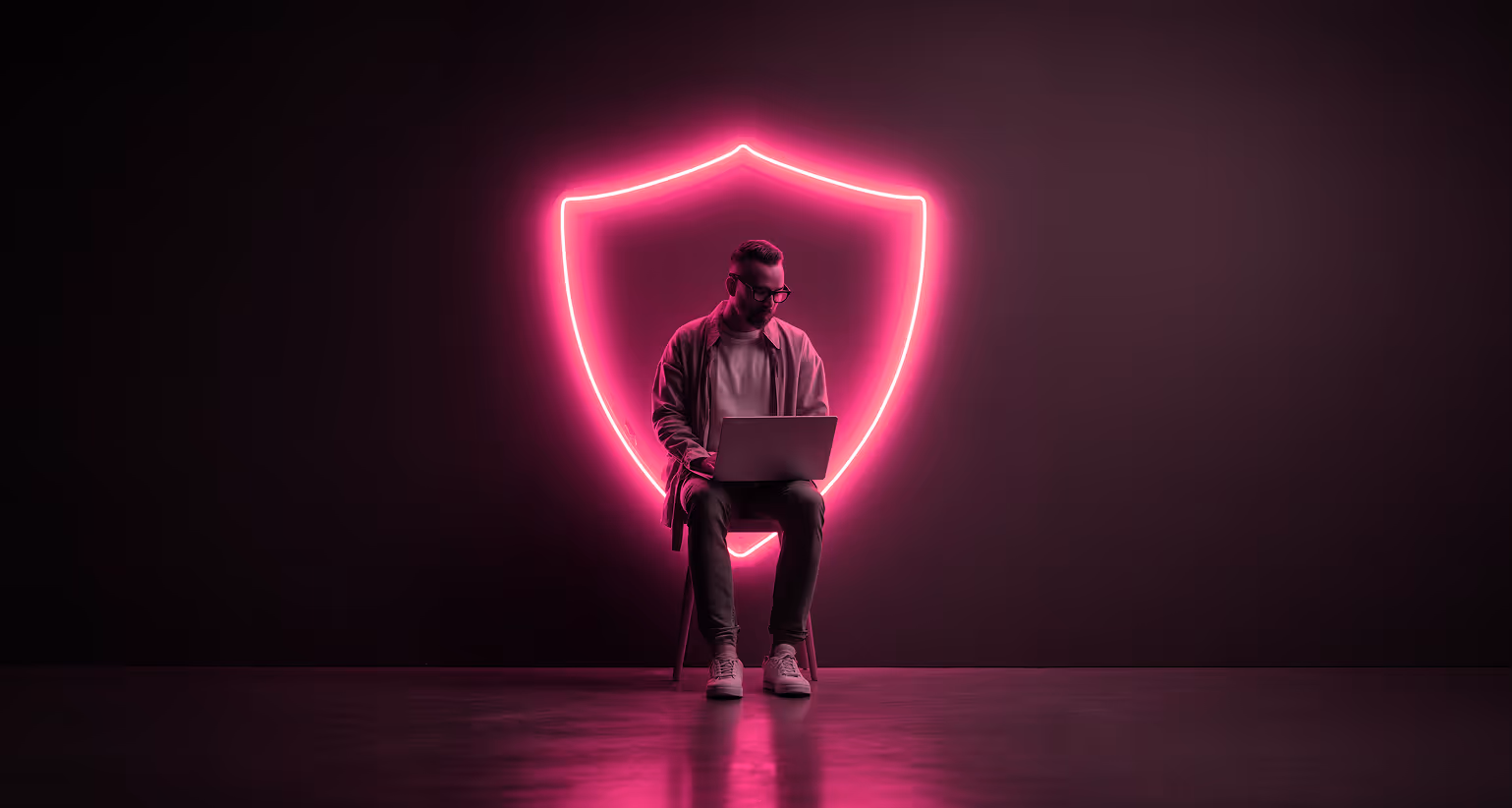 Man sitting on chair using laptop in front of a glowing pink neon shield on black background.