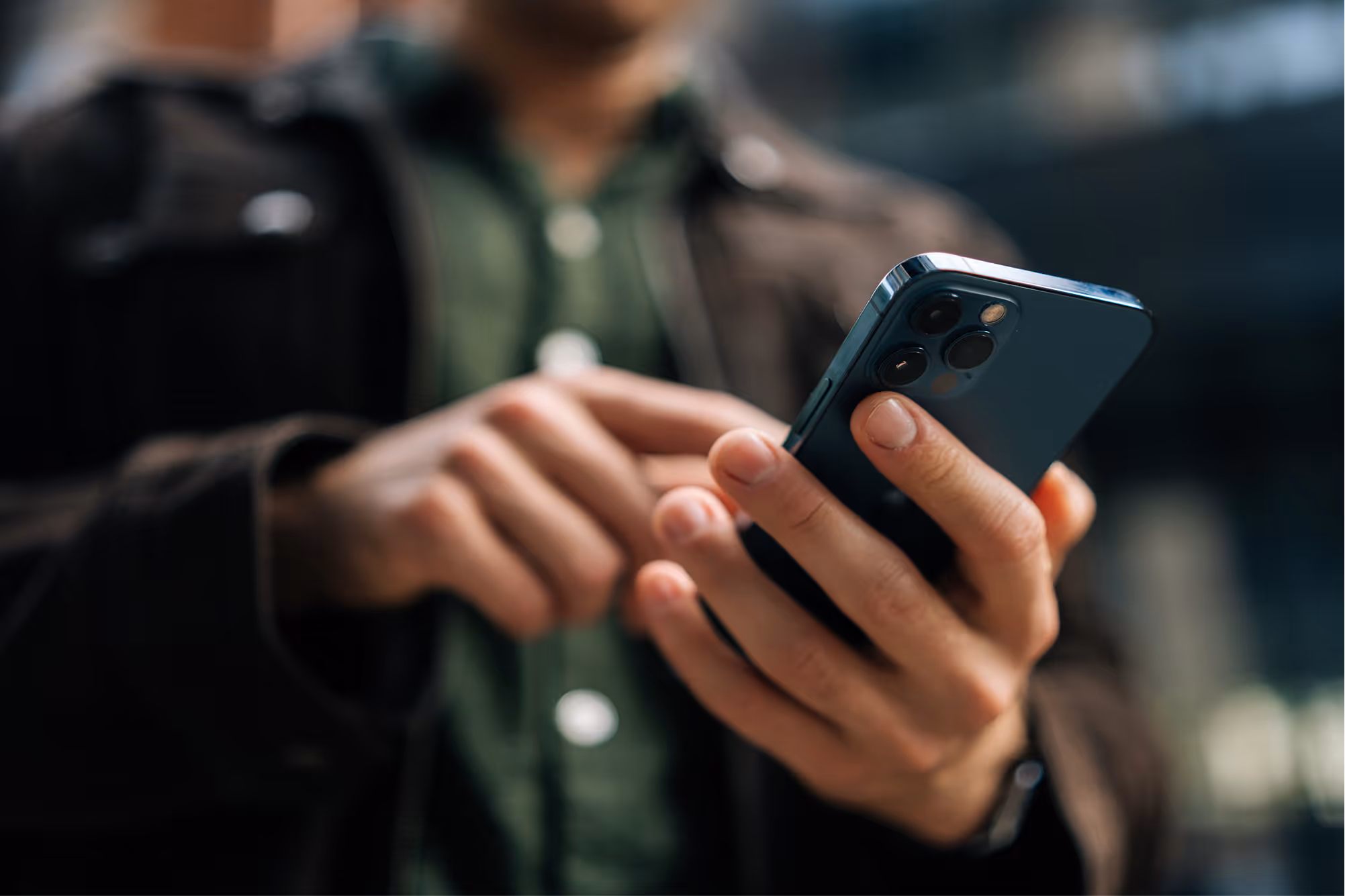 Person in brown jacket using a smartphone indoors, focus on hands and phone with triple camera lenses.