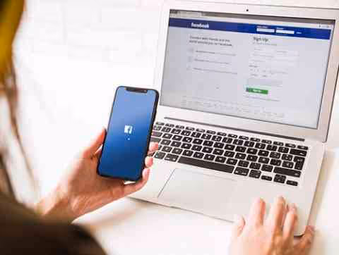 Person hält ein Smartphone mit Facebook-Logo vor einem Laptop, auf dem die Facebook-Anmeldeseite geöffnet ist.