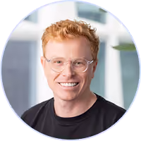 Smiling man with curly red hair and glasses wearing a black shirt in a modern office.