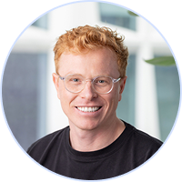 Smiling man with curly red hair and glasses wearing a black shirt in a modern office.