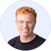 Smiling young man with curly red hair, glasses, and a black shirt on a light background.