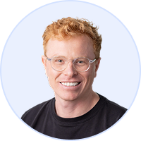 Smiling young man with curly red hair, glasses, and a black shirt on a light background.