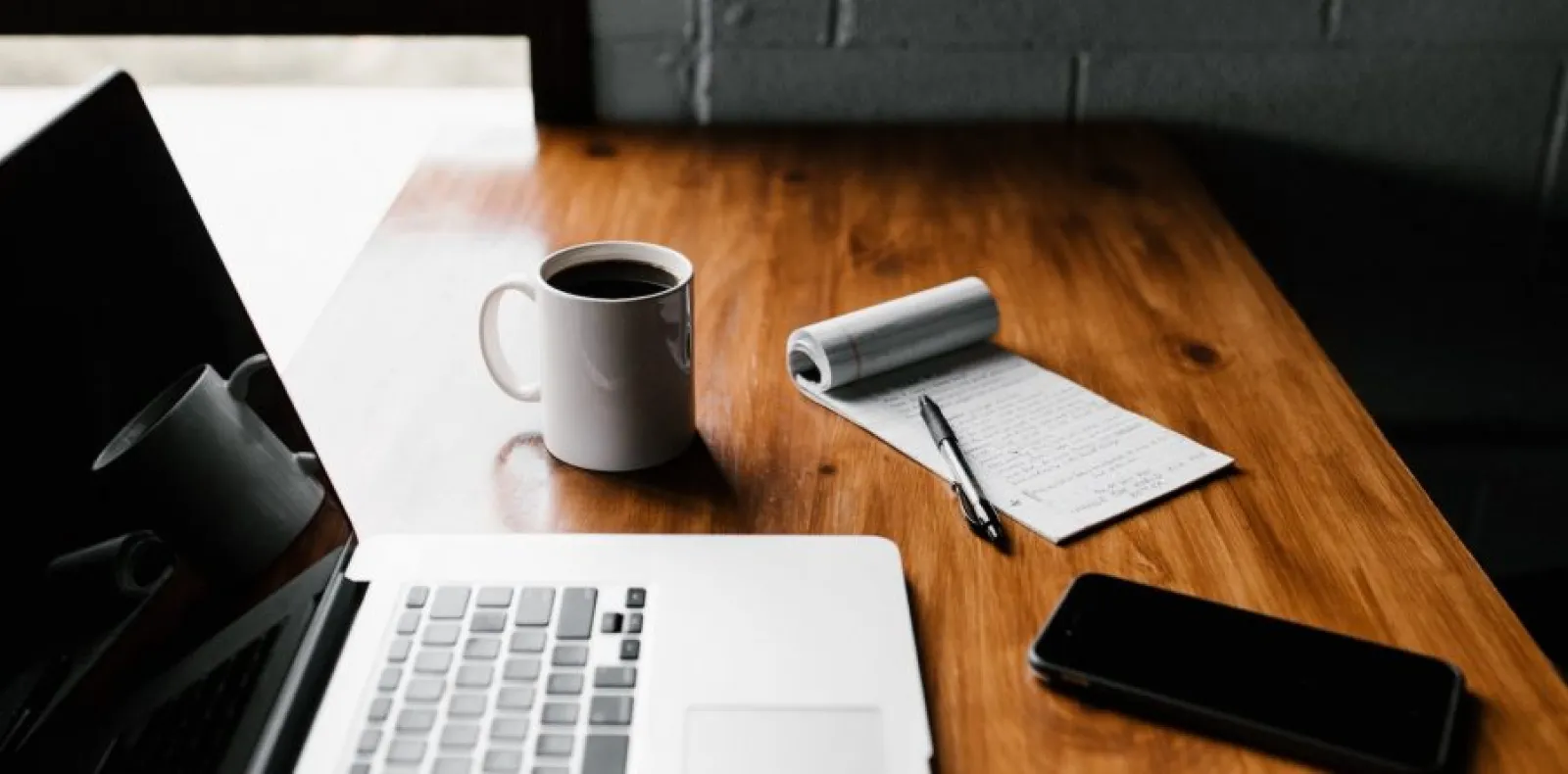Worth Recruiting Toronto. Wooden desk with a laptop, a white coffee mug filled with black coffee, a notepad with handwritten notes and a pen, and a smartphone.