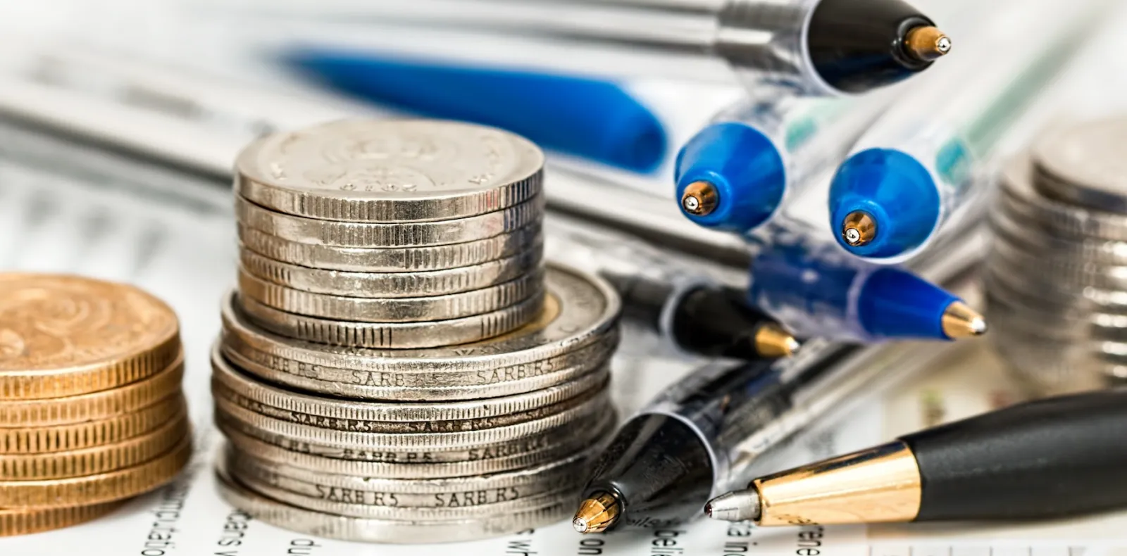 Worth Recruiting Toronto. Stacks of coins placed next to several ballpoint pens on a sheet of paper with printed text.