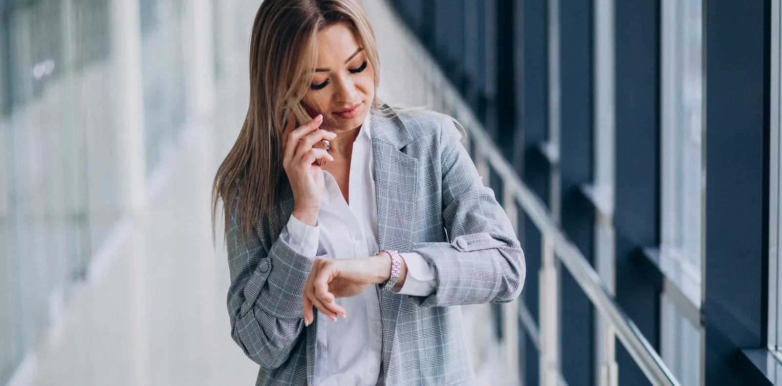 Worth Recruiting Toronto. Young business woman in a gray plaid blazer looking at her wristwatch while talking on a smartphone in a modern office hallway.