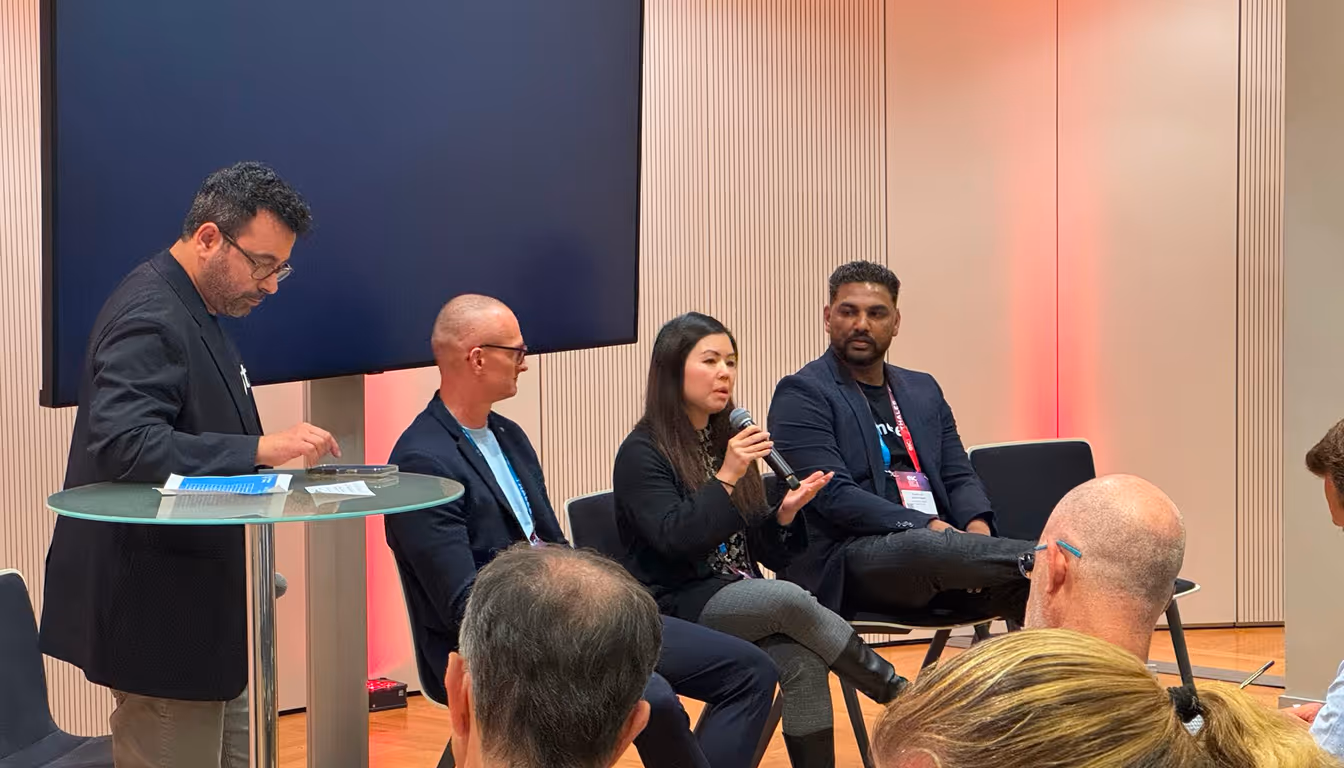 Panel discussion with four speakers seated, one woman speaking into a microphone while a man stands at a glass table nearby.