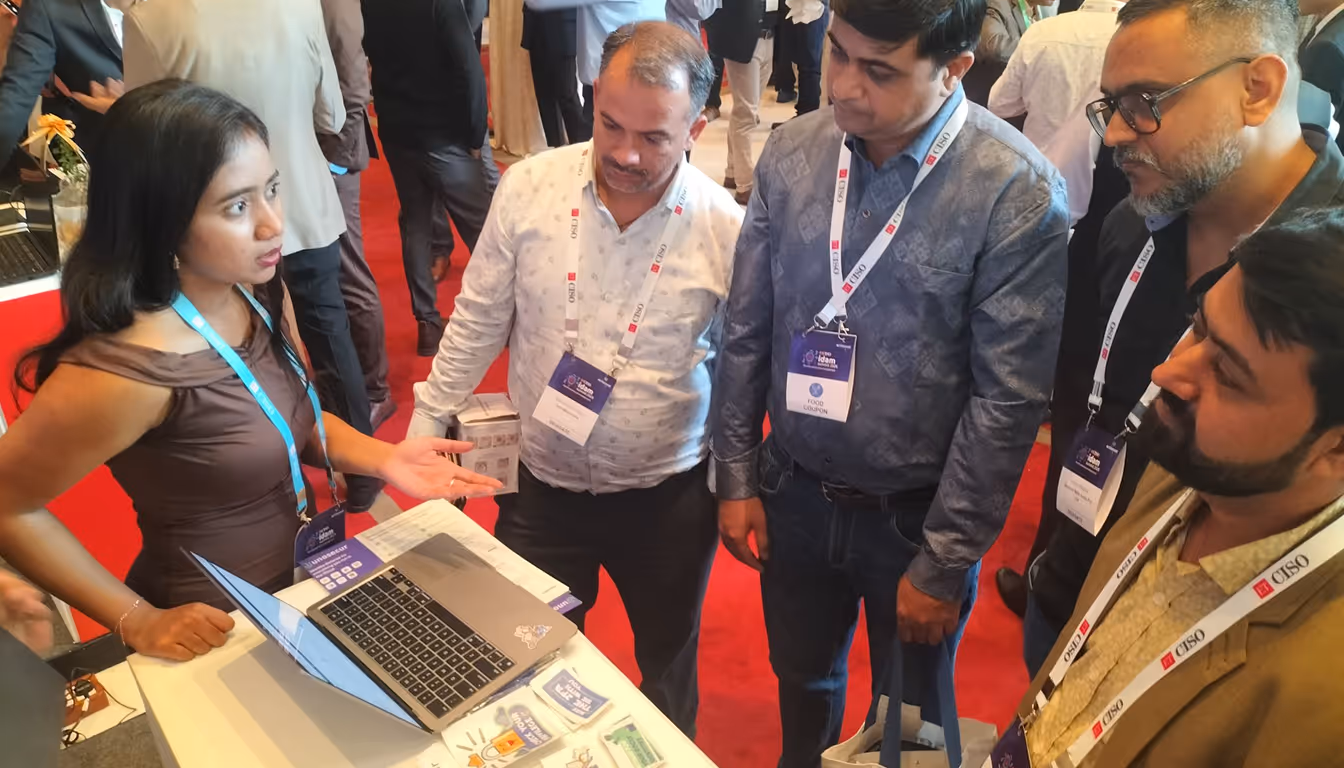 A woman explaining something on a laptop to four men wearing conference badges on a red carpeted floor.