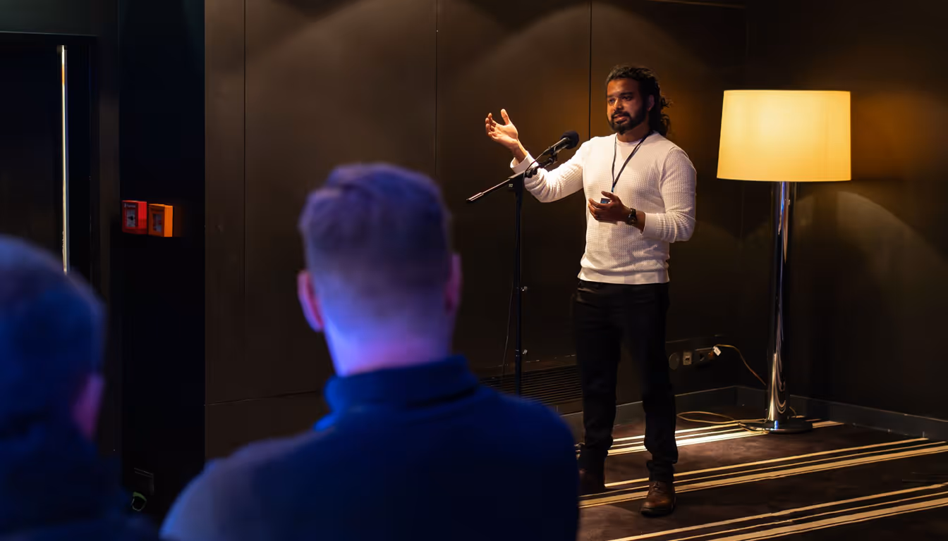 Man in a white sweater speaking into a microphone in front of an audience in a dim room with a yellow floor lamp.