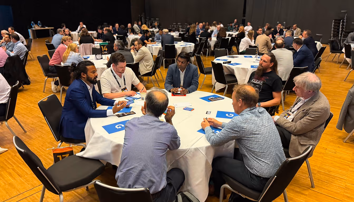 Group of men engaged in discussion around a round table in a conference setting with other tables and attendees in the background.