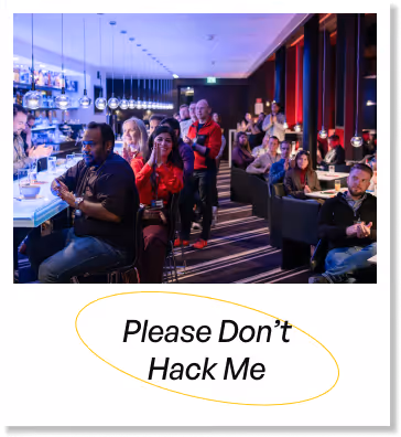People seated and standing in a modern lounge or bar setting, with a neon sign below saying 'Please Don't Hack Me'.