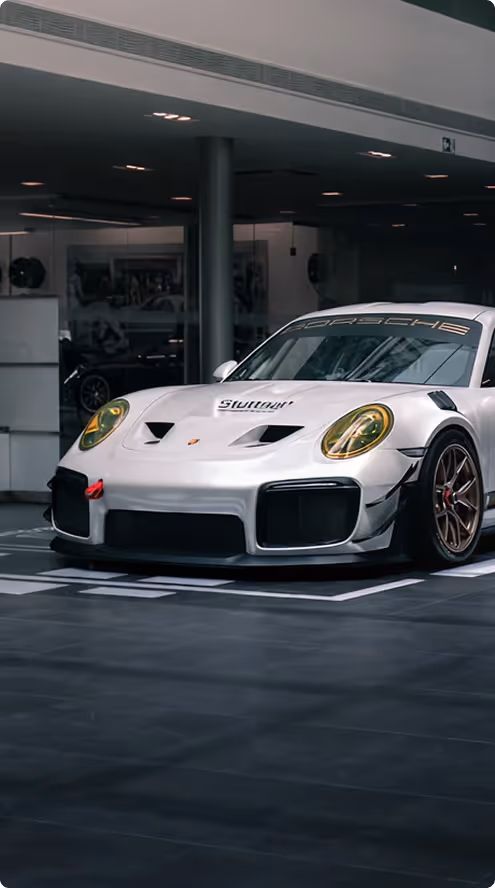 White Porsche race car with yellow-tinted headlights parked indoors in a showroom.